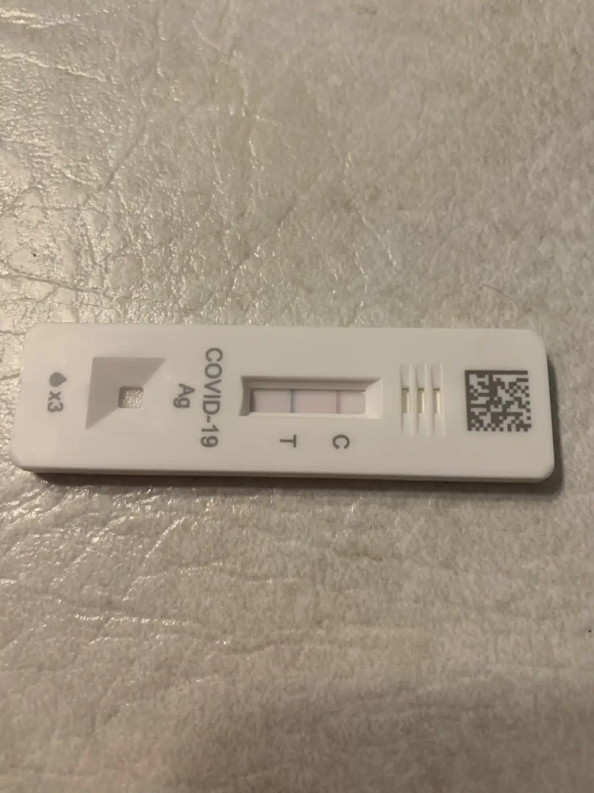 A white COVID-19 antigen test kit rests on a textured, light-colored surface. The test window shows two lines, one at the…