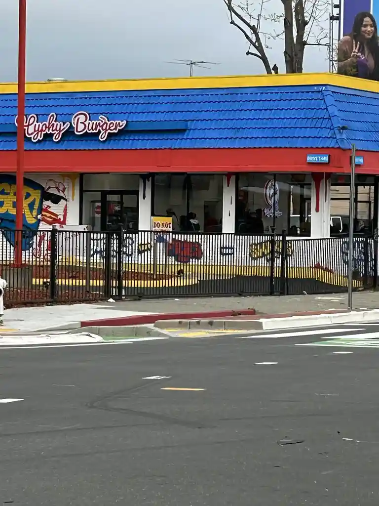An exterior shot of a brightly colored 'Lyphy Burger' restaurant with a blue roof, red trim, and a black fence in front. The…