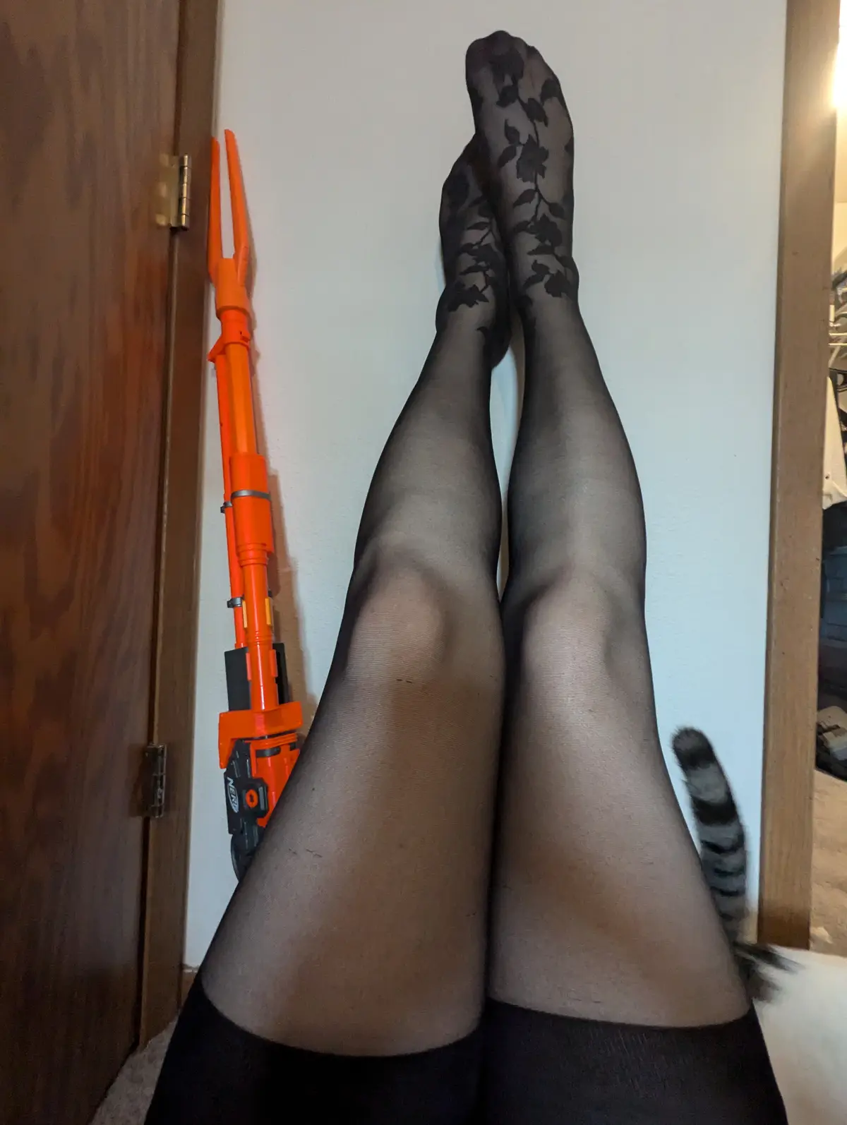 A person wearing black floral-patterned tights has their legs stretched upward against a white wall, with a bright orange…