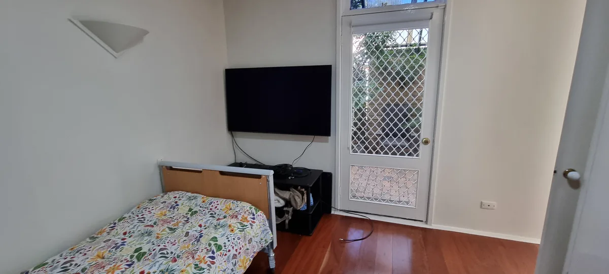 A simple bedroom with a bed covered in a colorful floral blanket, a wall-mounted TV, and a door with a security screen. The…