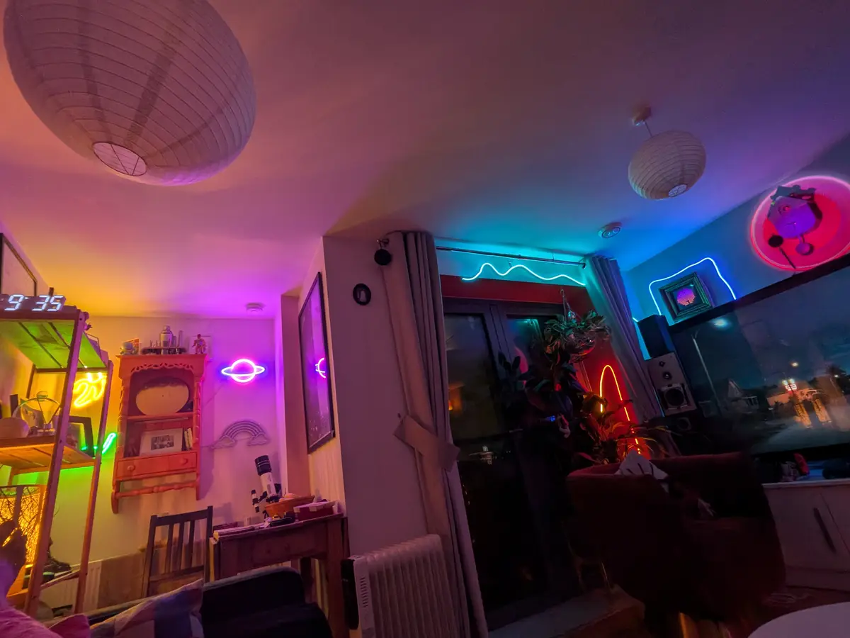 A dimly lit room bathed in colorful neon lights, featuring paper lanterns, a digital clock showing 9:35, a Saturn neon sign…