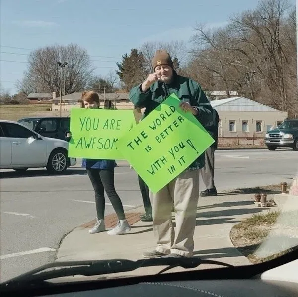 Two people stand on a sidewalk holding bright green signs. The person in front holds a sign reading 'THE WORLD IS BETTER…