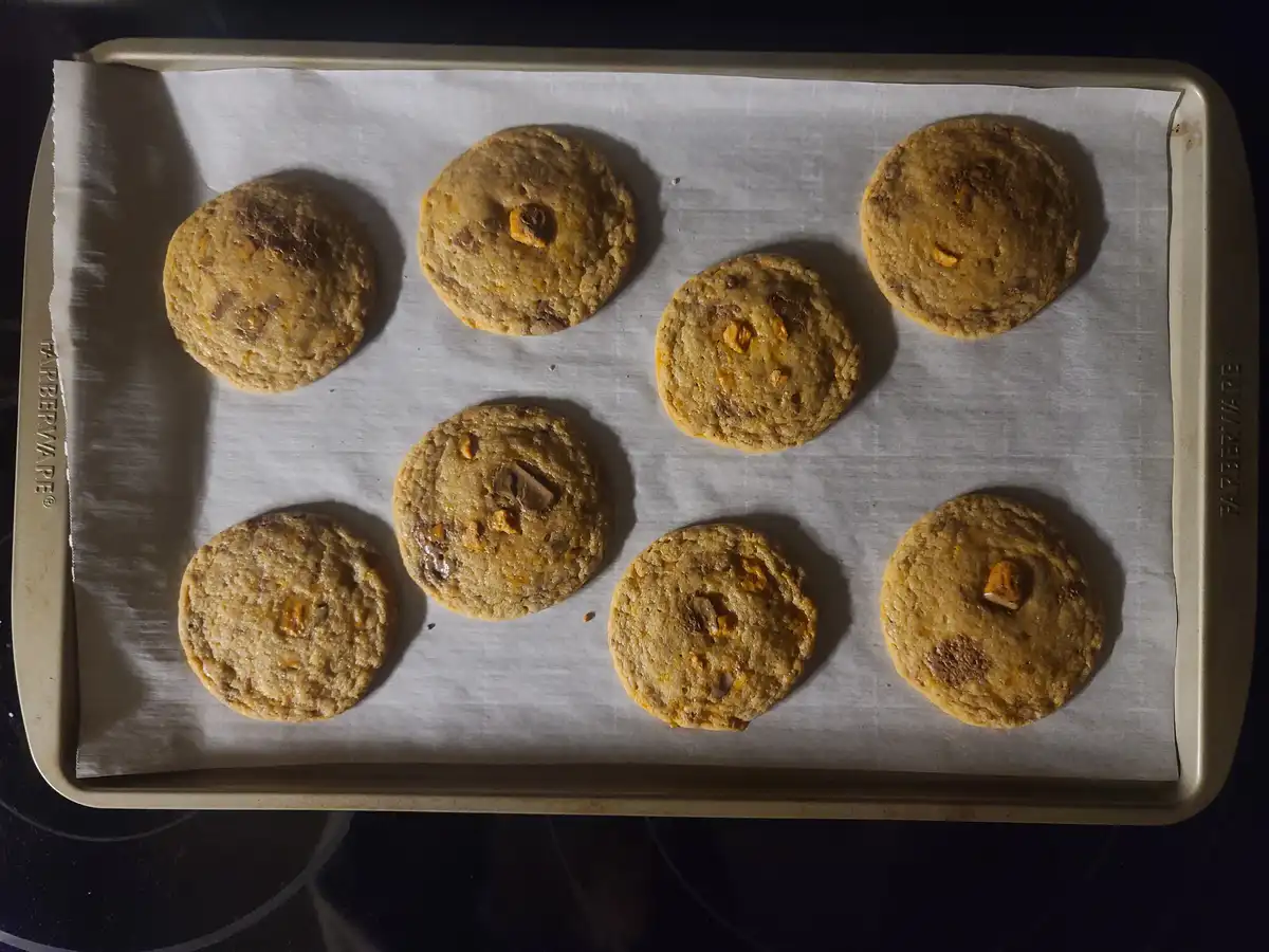 A baking sheet lined with parchment paper holds eight freshly baked chocolate chip cookies, with one cookie noticeably…