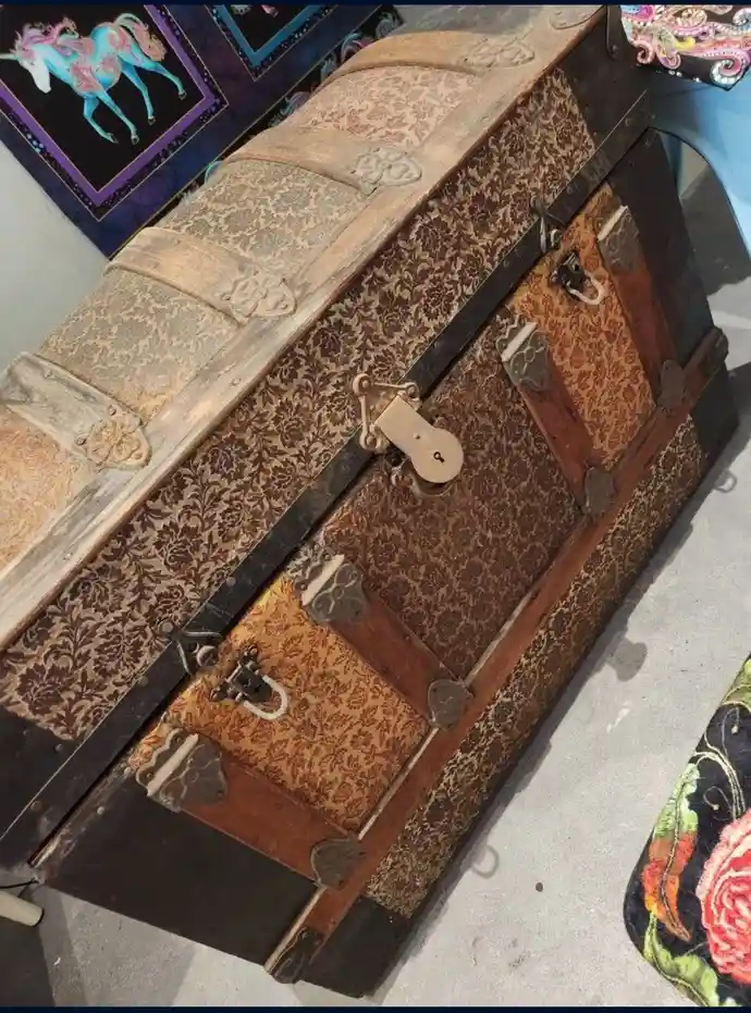 a large, ornate, vintage wooden trunk with floral-patterned fabric panels and metal clasps is shown from an angled…