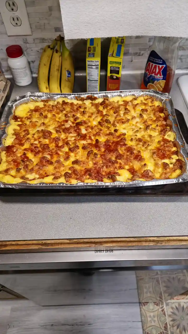 A freshly baked, cheesy casserole sits in a foil pan on a kitchen counter, with bananas, cereal boxes, and Ajax cleaner…