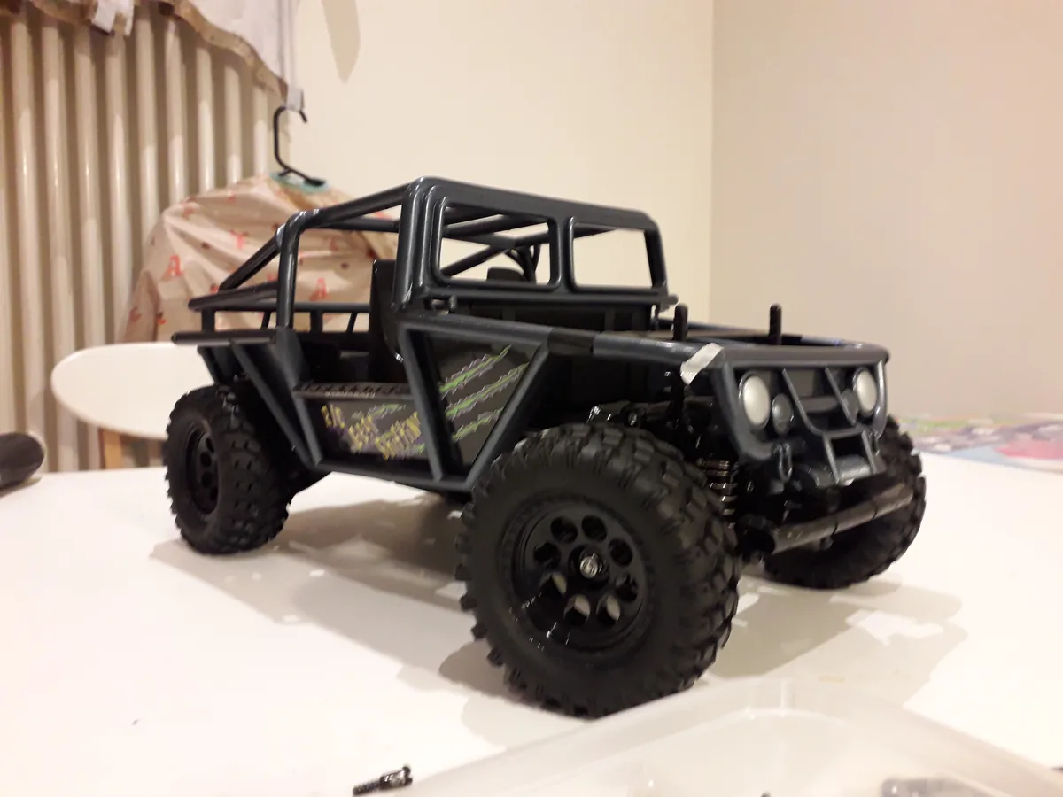 A black remote-controlled off-road vehicle with large knobby tires and a roll cage sits on a white surface, featuring green…