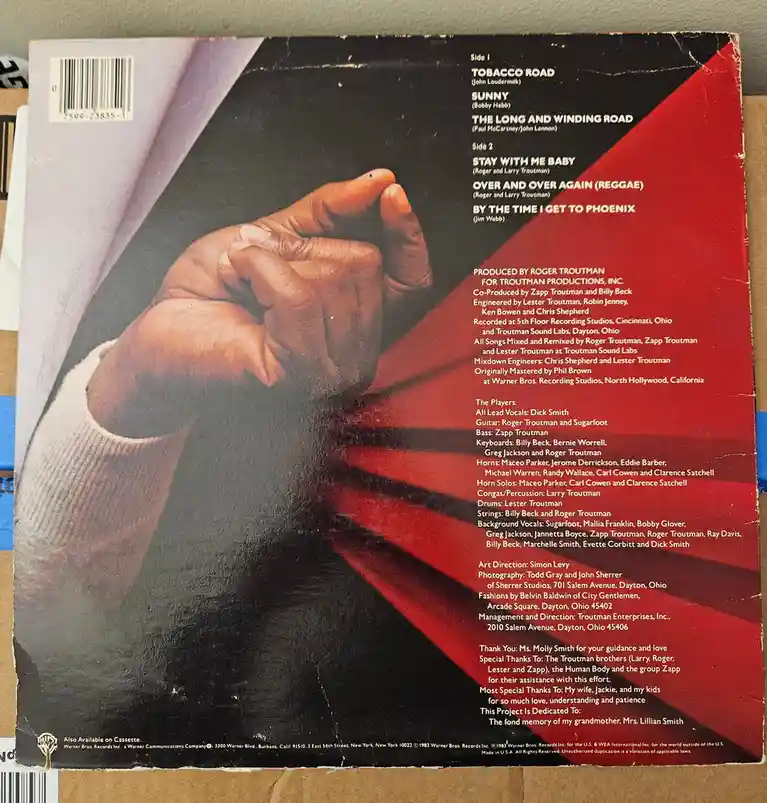The image shows the back cover of a 1983 Warner Bros. vinyl record album featuring a close-up of two clasped hands against a…