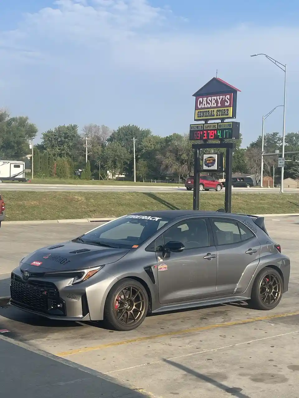 A heavily modified gray Toyota Corolla GR is parked in front of a Casey's General Store gas station. The digital sign behind…