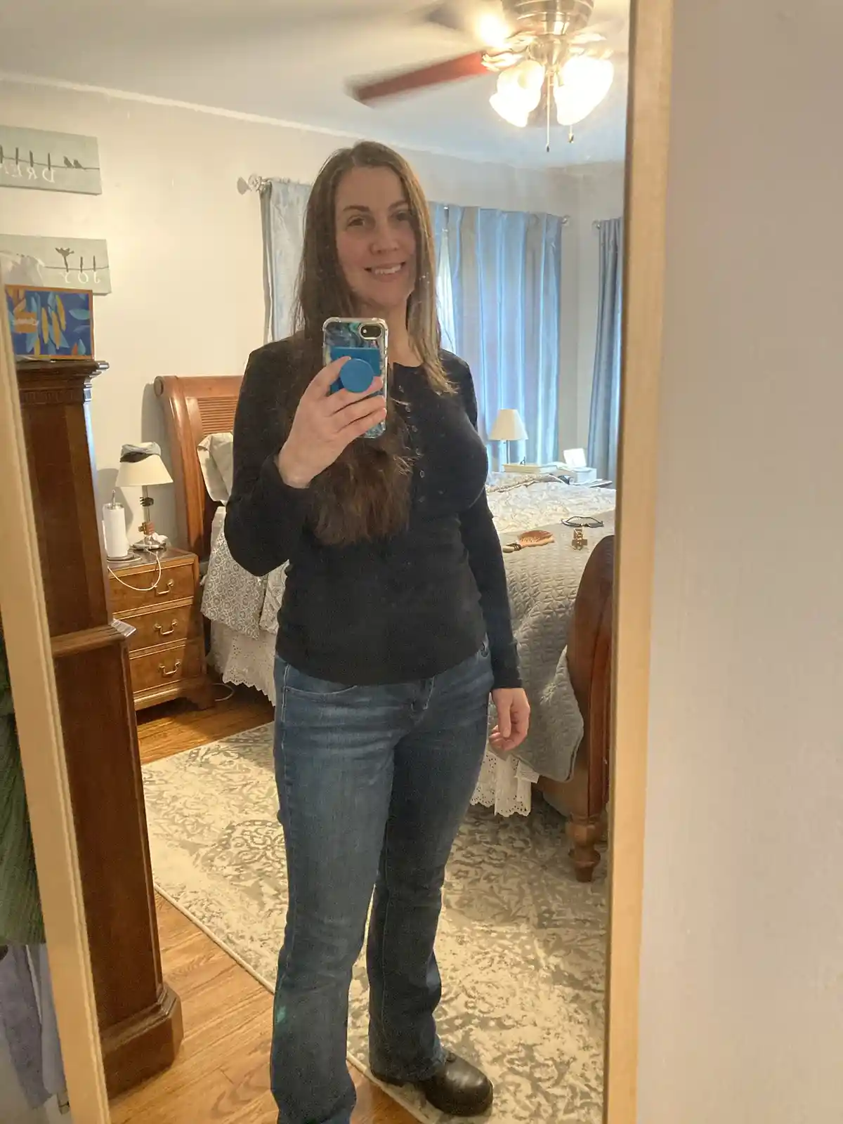 A woman takes a mirror selfie in her bedroom wearing a black long-sleeved shirt and blue jeans, smiling at the camera while…