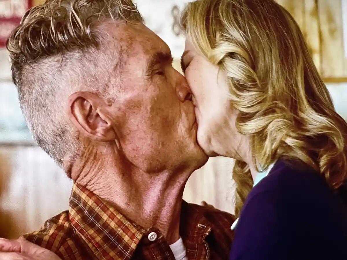 An older man with gray hair and a plaid shirt is kissing an older woman with blonde hair on the cheek. The image has text…