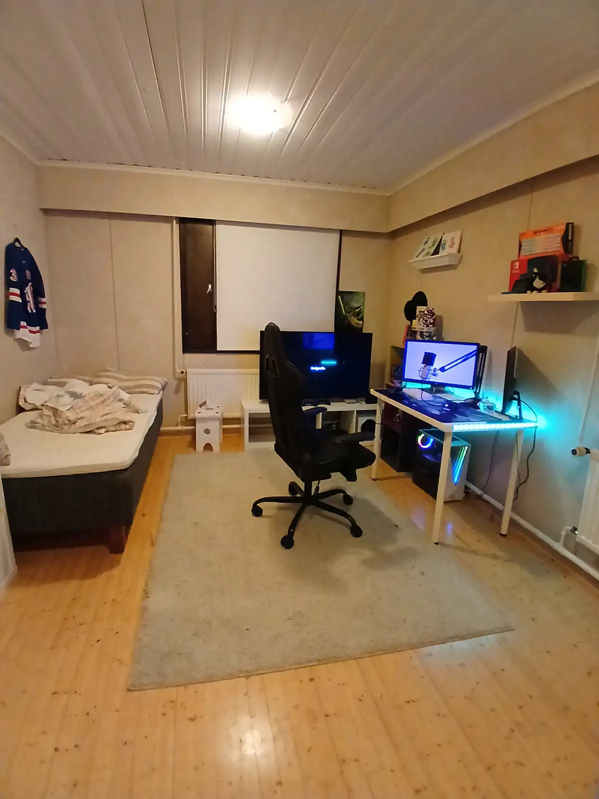 A room with a bed on the left, a gaming chair, and a desk with a glowing computer setup on the right. A jersey hangs on the…