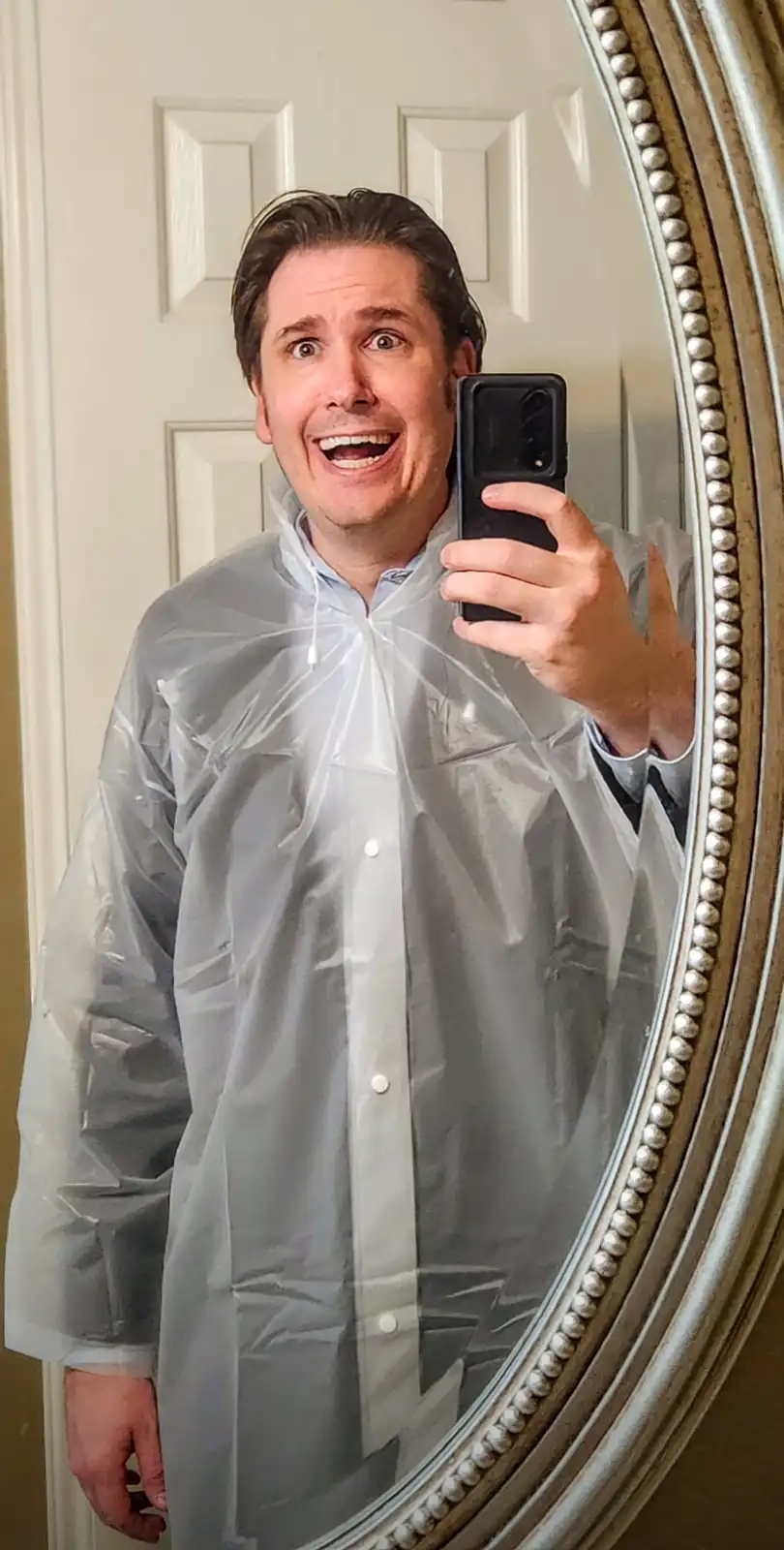 A man takes a mirror selfie wearing a transparent plastic raincoat over a button-up shirt, looking extremely excited and…