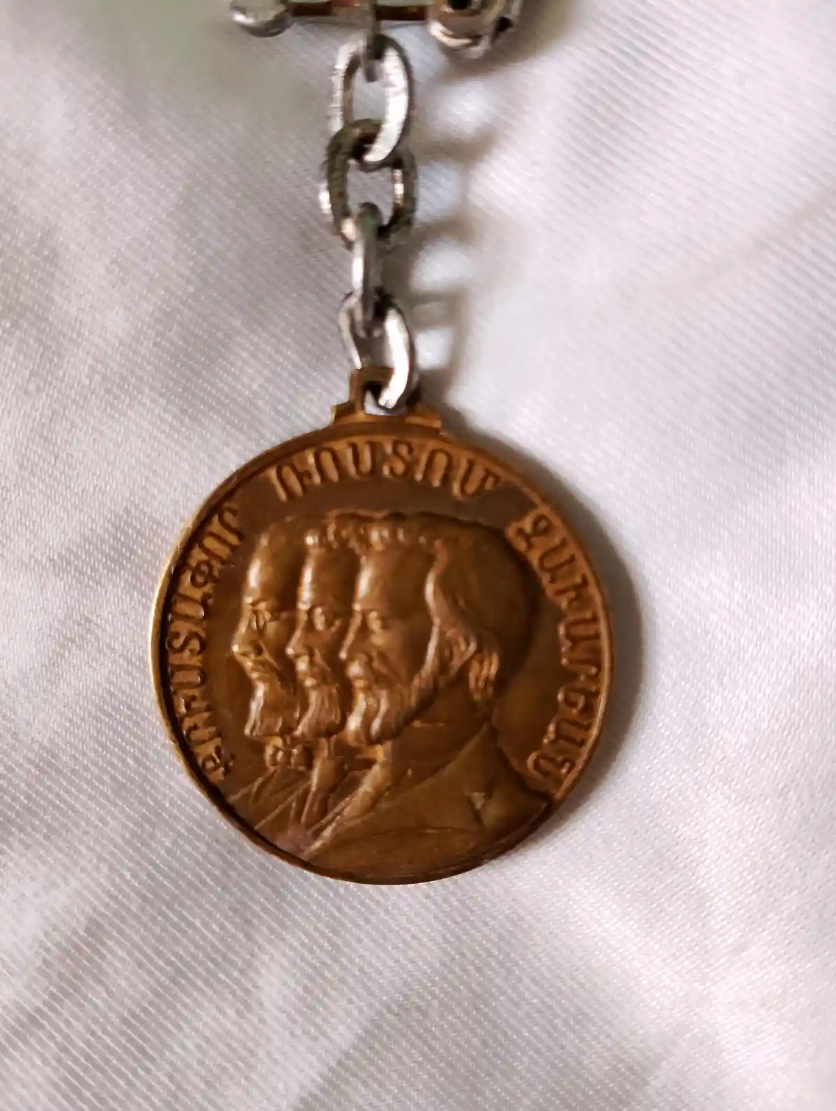 A close-up photo shows a bronze religious medal hanging from a silver chain, resting on a white textured fabric. The medal…