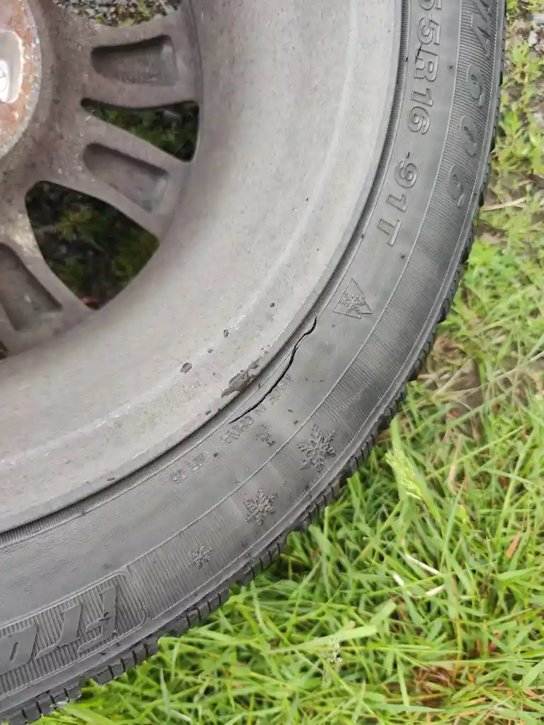 A close-up photo shows a worn winter tire resting on green grass, with visible snowflake symbols and a prominent crack…