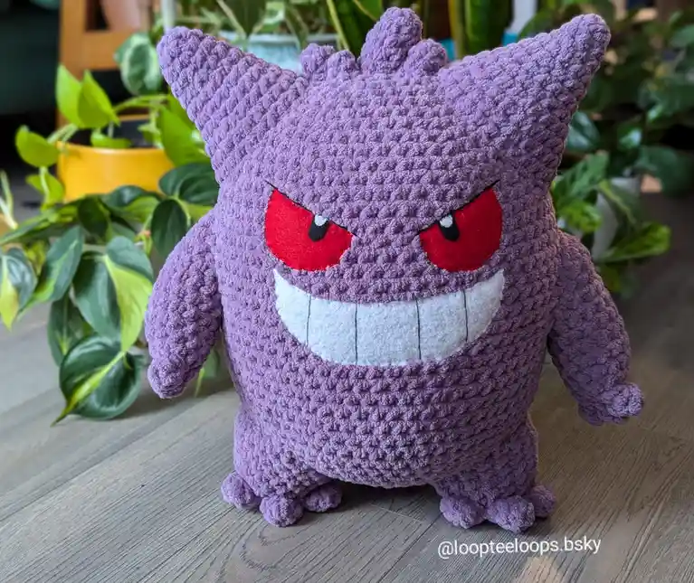 A handmade, purple crocheted plush toy of the Pokémon Gengar, featuring red eyes and a wide white grin, stands on a wooden…