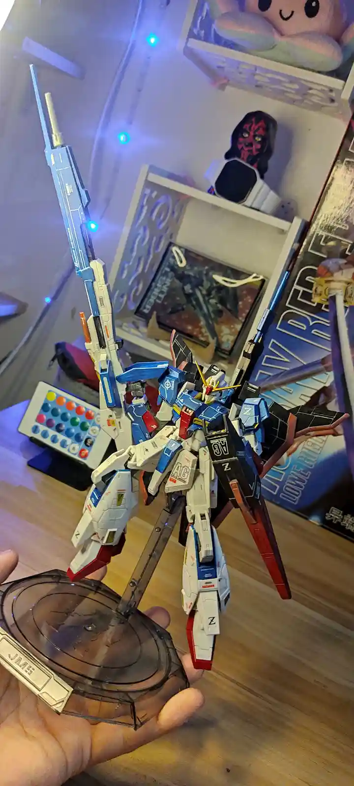 A person holds a detailed blue, white, and red Gundam model kit on a clear plastic stand, with a large blue and silver…