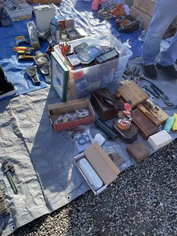 An outdoor yard sale display on a blue tarp spread over gravel, featuring a large clear plastic bin overflowing with…