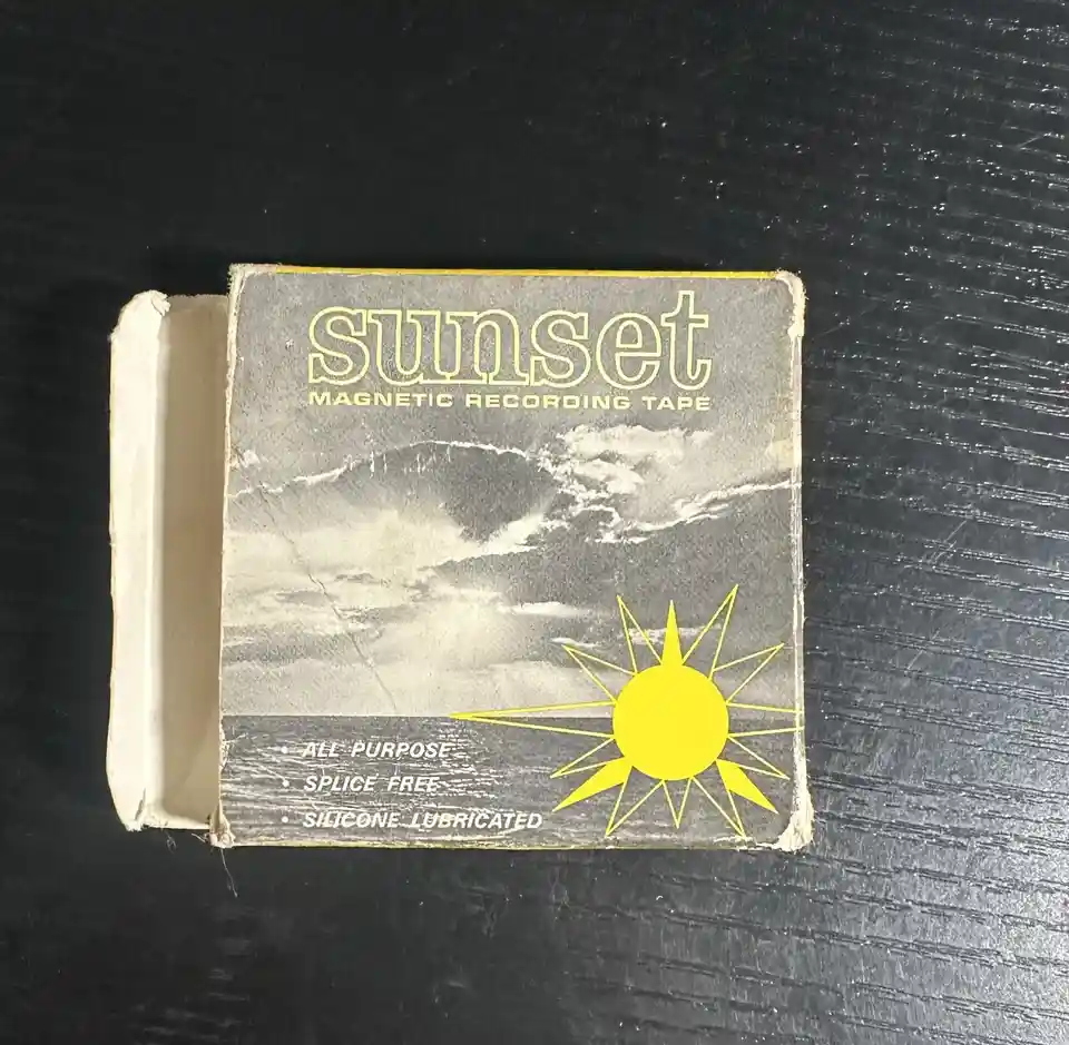 A vintage, weathered box of 'Sunset Magnetic Recording Tape' sits on a dark surface. The box features a black-and-white…