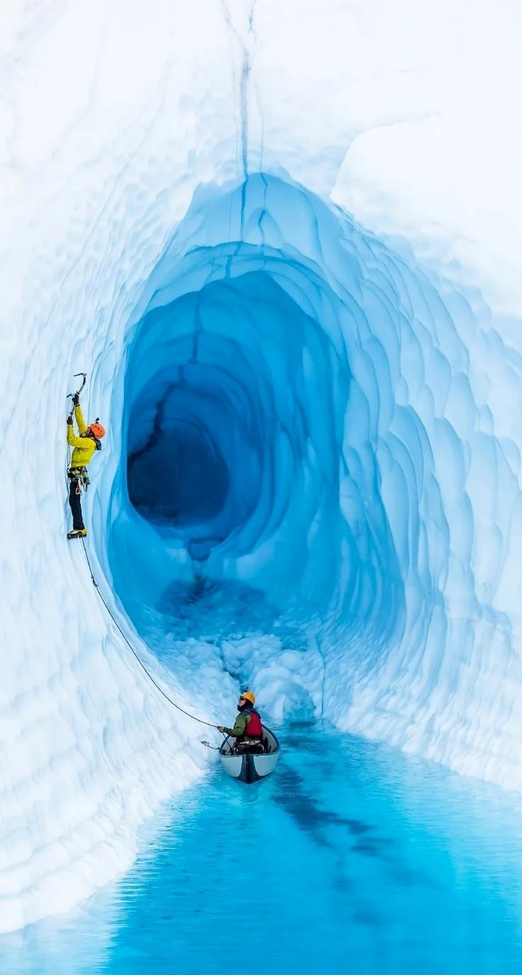 Two people are inside a massive, blue ice cave. One person in a yellow jacket and orange helmet is ice climbing up the left…