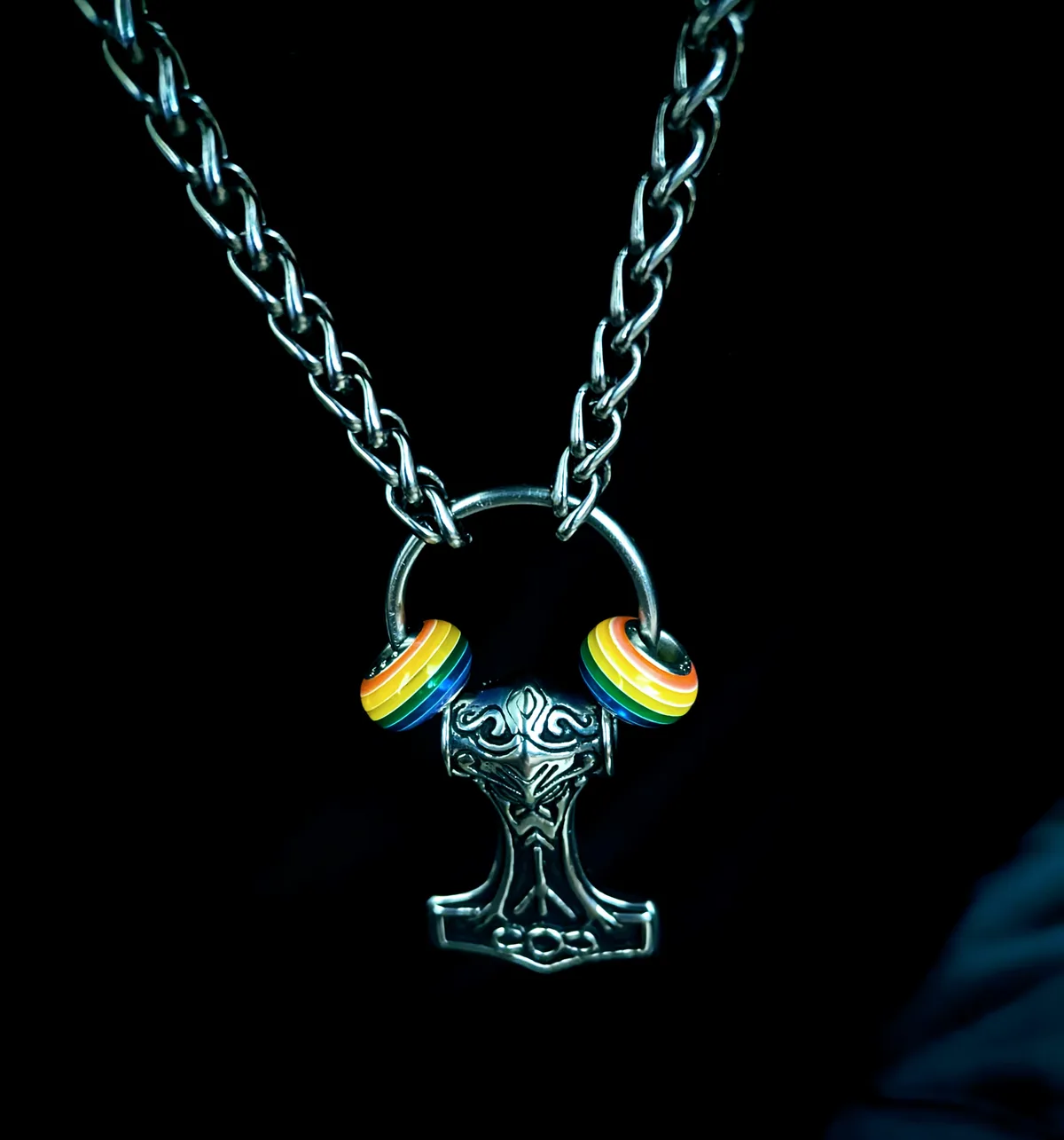 A silver Thor's hammer pendant hangs from a chain against a black background, with rainbow-colored headphone earcups…