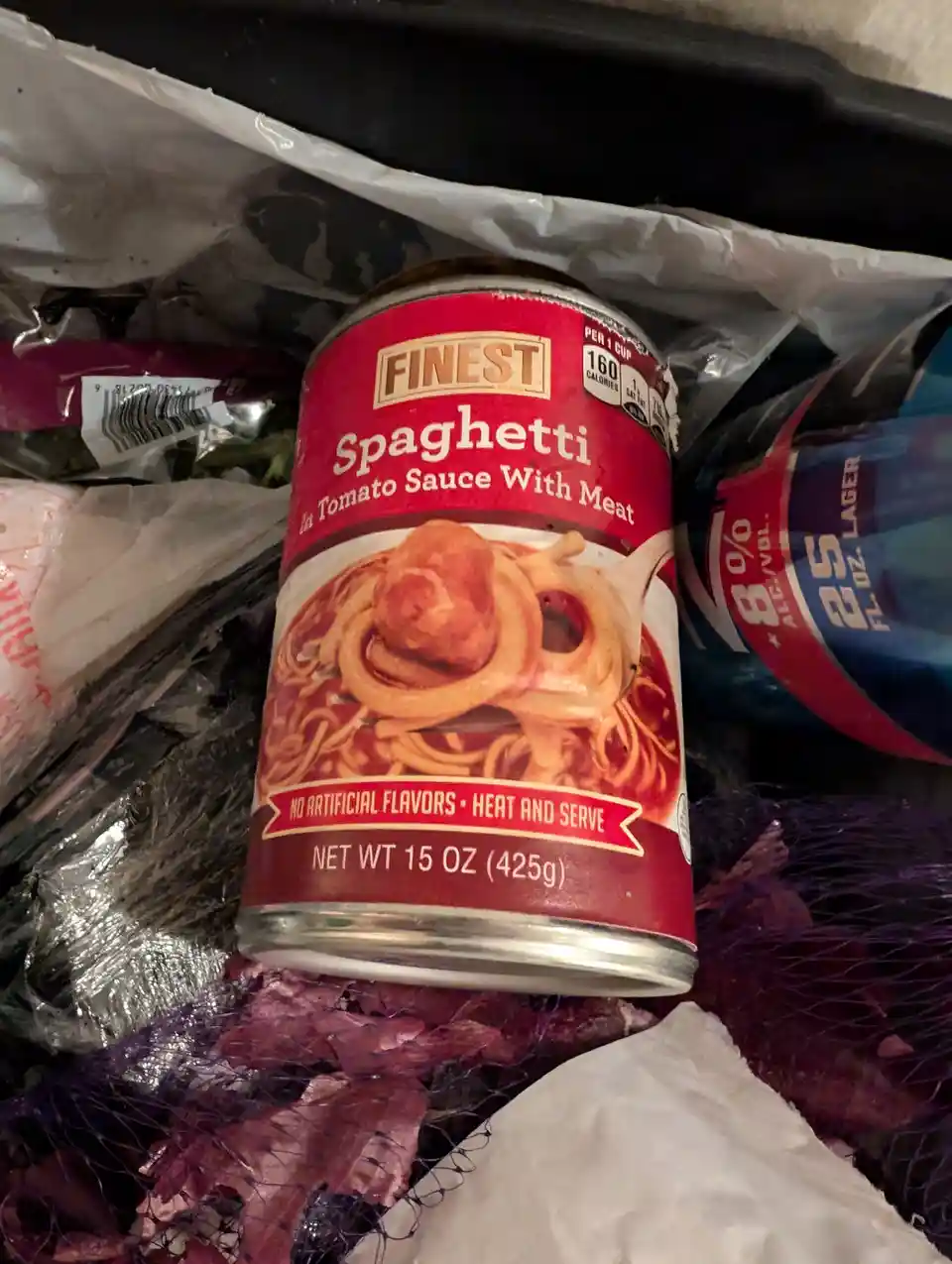 A can of 'Finest' brand spaghetti in tomato sauce with meat sits among grocery bags and produce, its label showing a picture…