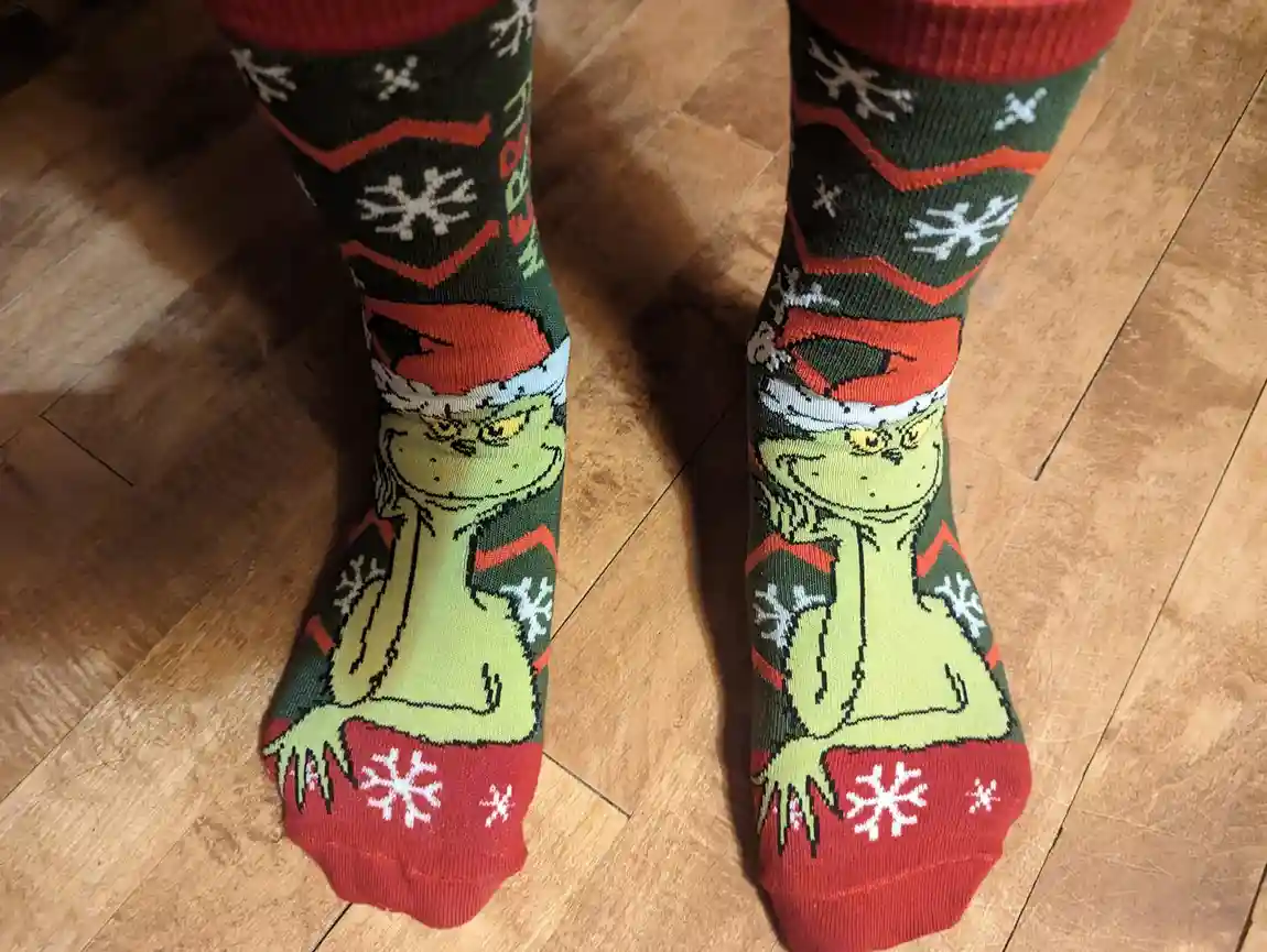 A close-up photo shows a person's feet wearing festive green and red Christmas socks featuring the Grinch character wearing…
