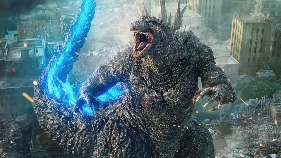 A giant, dark-scaled Godzilla stands amidst a ruined cityscape, roaring with his mouth wide open. His right arm is raised…