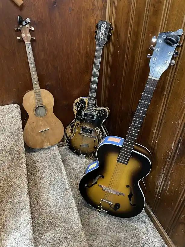Three guitars lean against a wooden wall on a carpeted staircase. The left guitar is a light-colored acoustic, the middle…