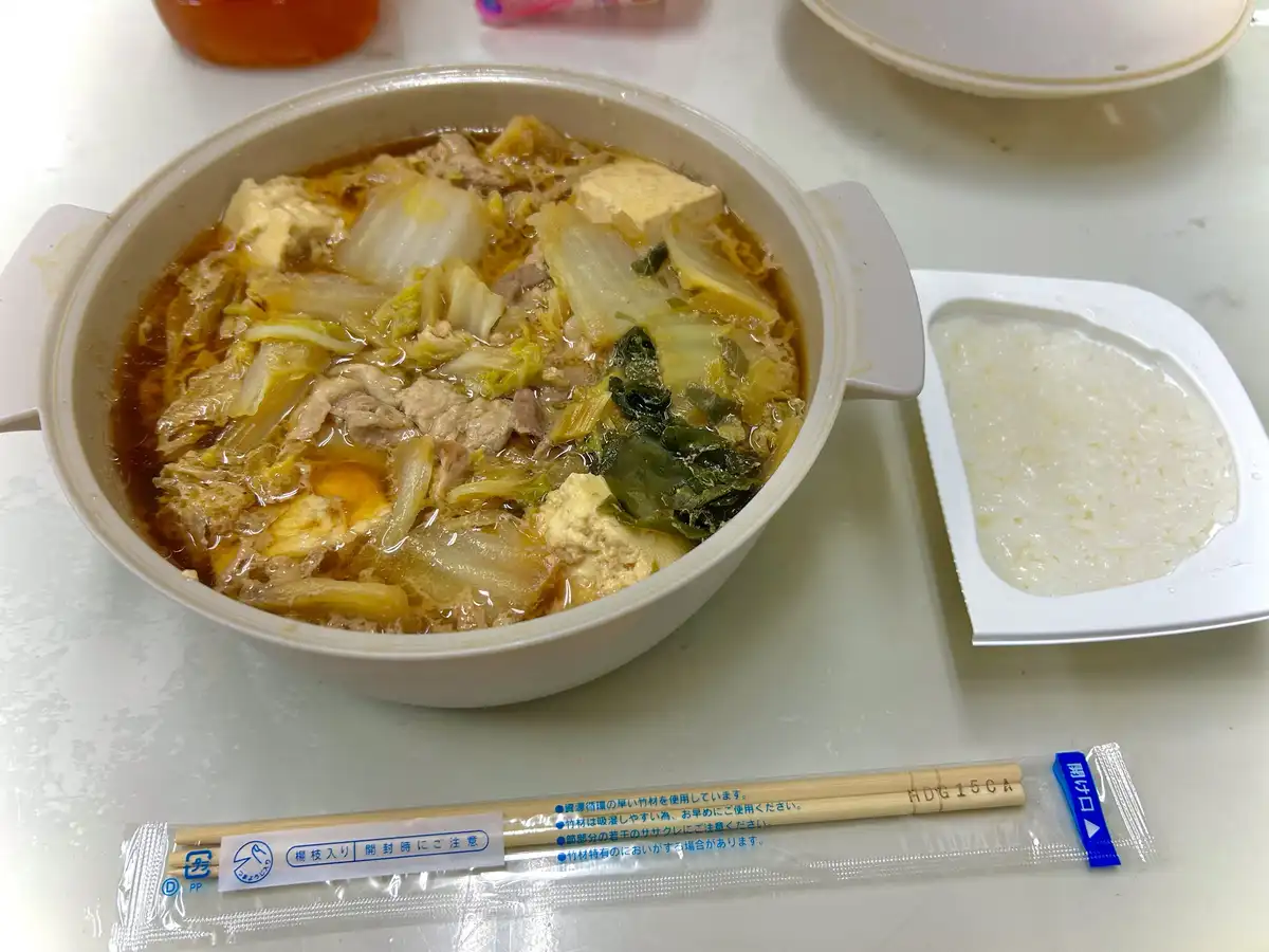 A close-up photo of a Japanese-style meal featuring a white plastic bowl filled with a hot, brown broth containing tofu…