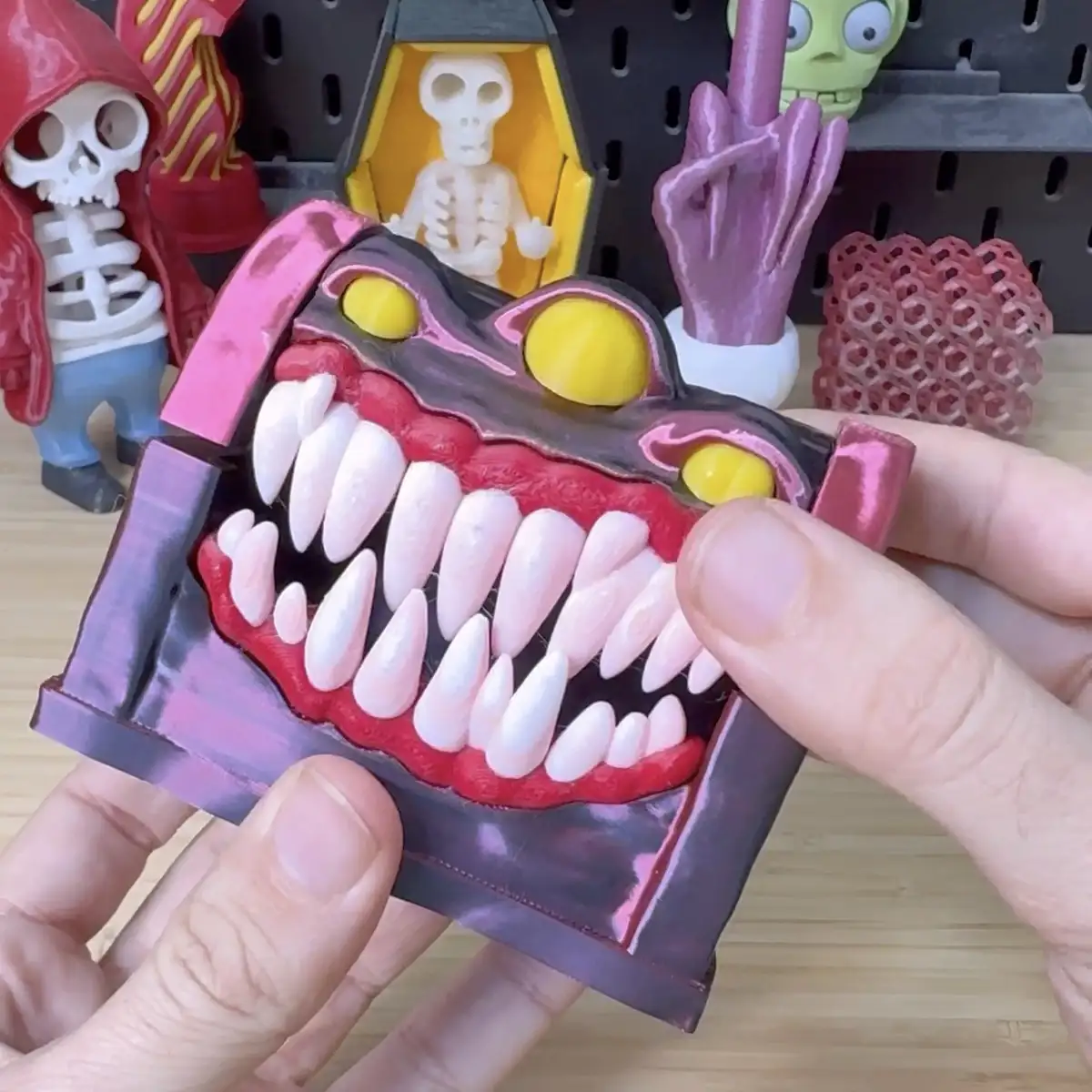 A person holds a 3D-printed, cartoonish monster head with a wide, toothy grin and yellow eyes. The monster's mouth is filled…