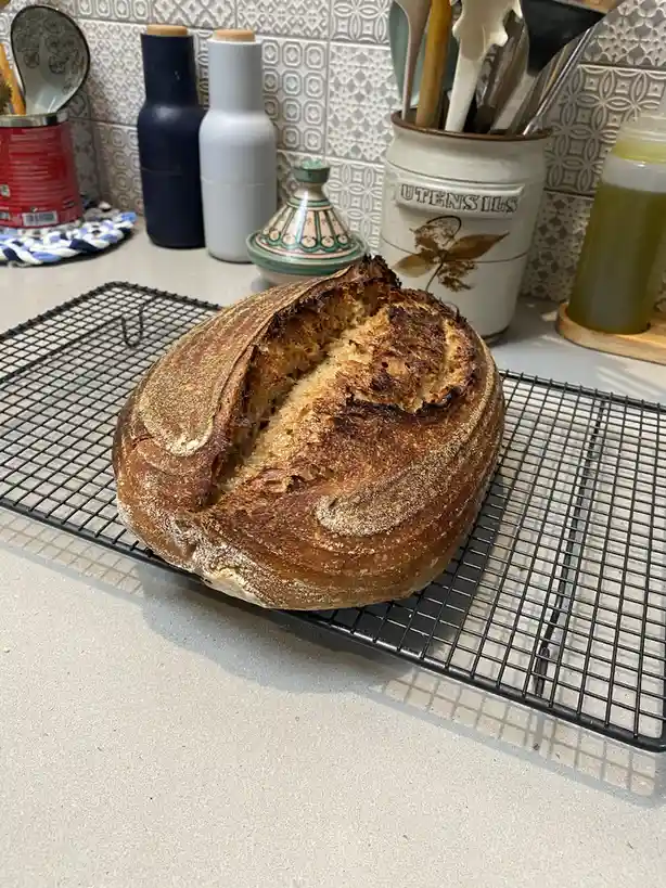 A round, rustic sourdough loaf with a deeply cracked, unevenly browned crust sits on a black wire cooling rack on a light…