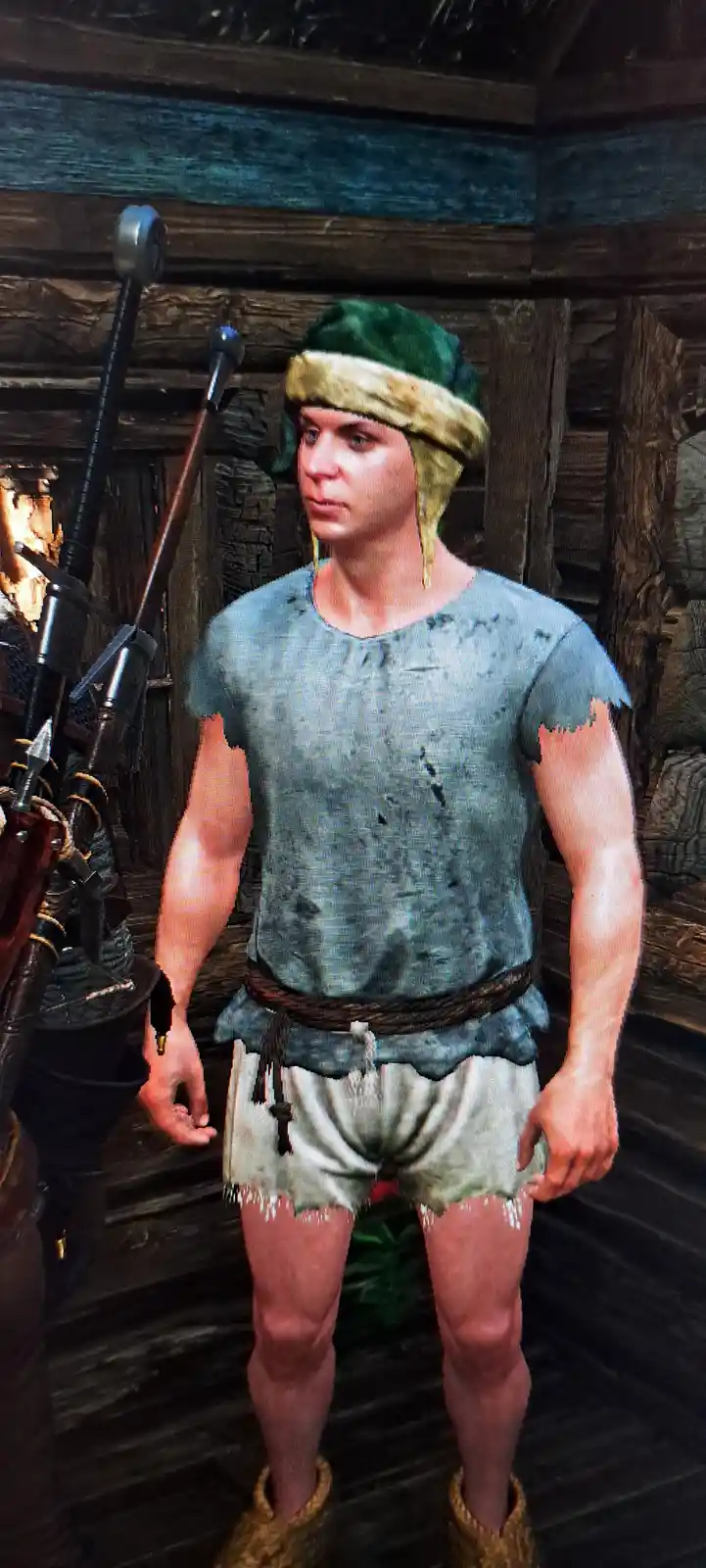 A video game character, resembling a medieval or fantasy warrior, stands in a wooden room with weapons visible nearby. He…