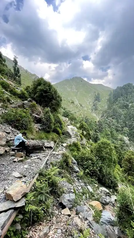 A person in a light blue shirt and dark pants sits on a low stone wall along a rugged, rocky mountain trail. The trail winds…