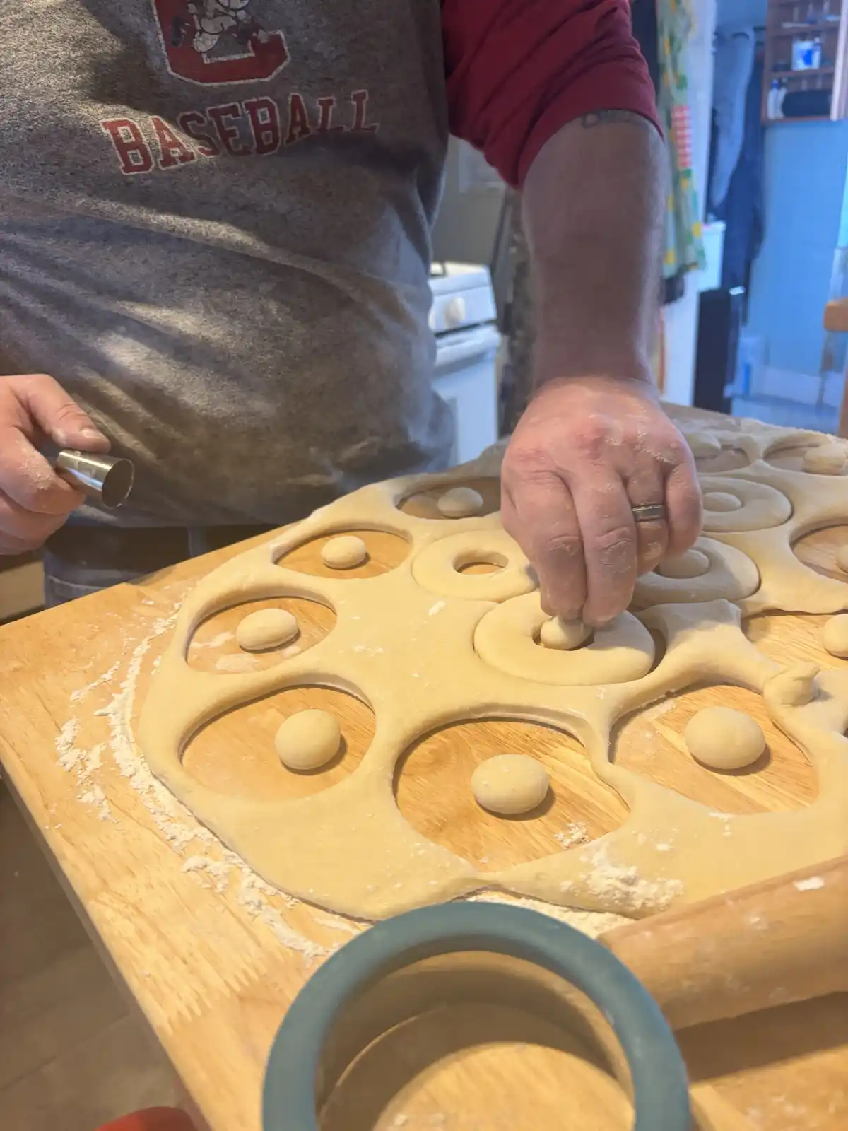 A person wearing a gray 'BASEBALL' apron over a red shirt is cutting dough on a wooden board using a round cutter, placing…