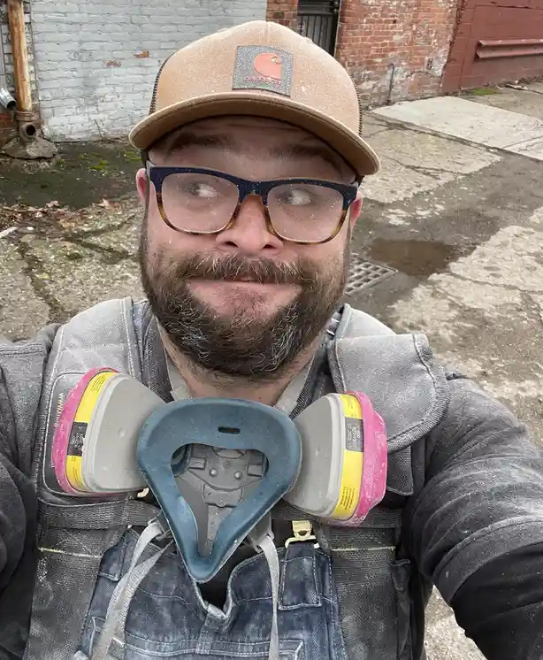A man with a beard and glasses, wearing a Carhartt cap and work overalls, takes a selfie outdoors. He is holding a…