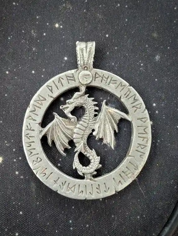 A silver dragon pendant with a circular border inscribed with reversed-looking runes, set against a dark speckled…