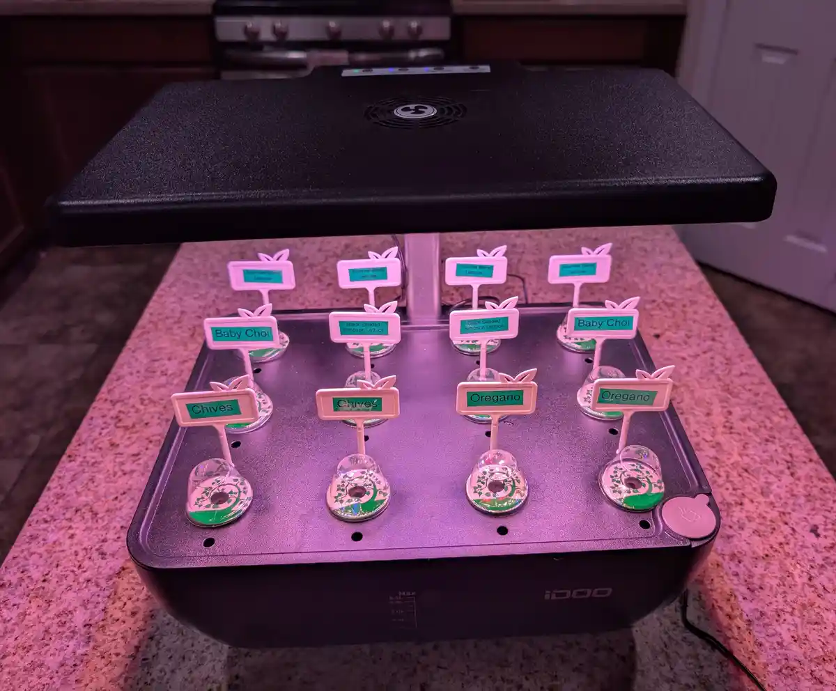 A black indoor herb garden device with pinkish-purple grow lights illuminates eight labeled plant pods on a kitchen counter…
