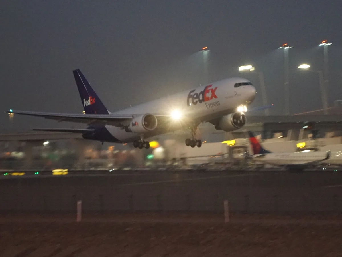 A blurry nighttime photo shows a FedEx Express cargo plane taking off or landing, its landing lights shining brightly…