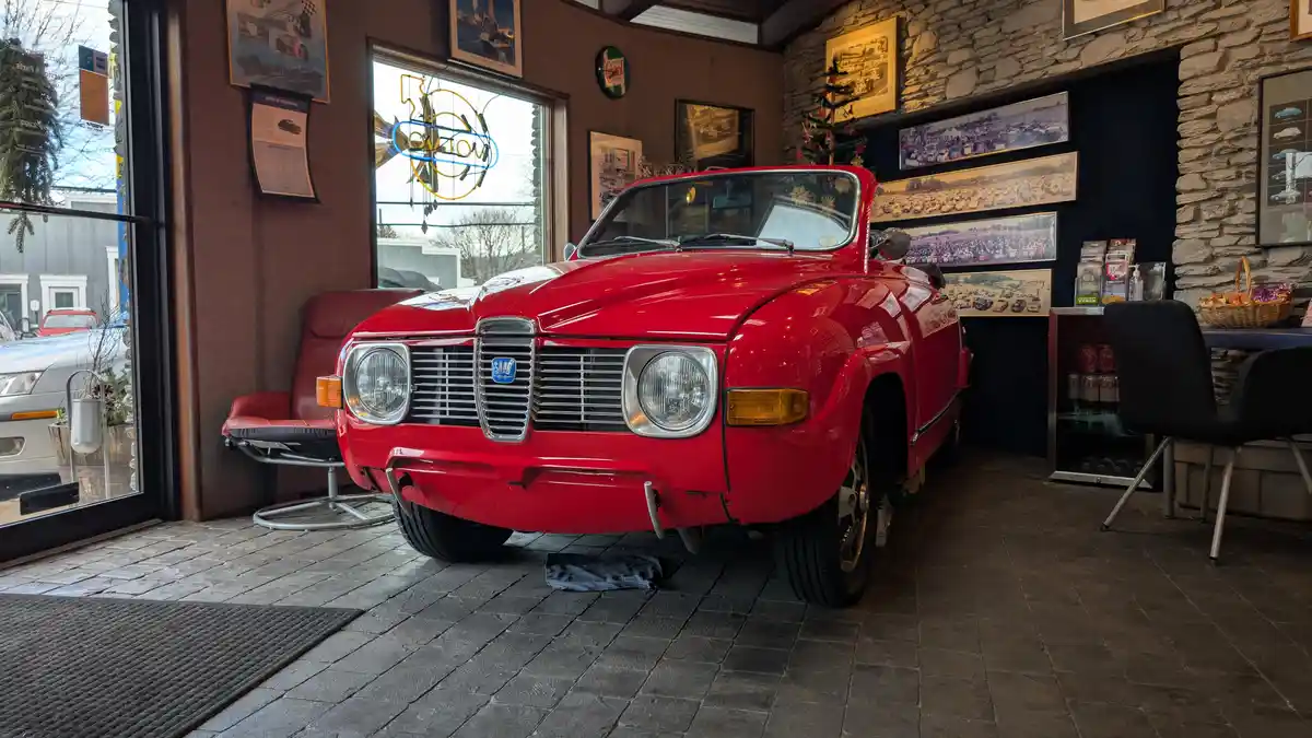 A red classic Volvo P1800 is parked indoors on a stone floor, with a 'volvo' neon sign visible through a window behind it…