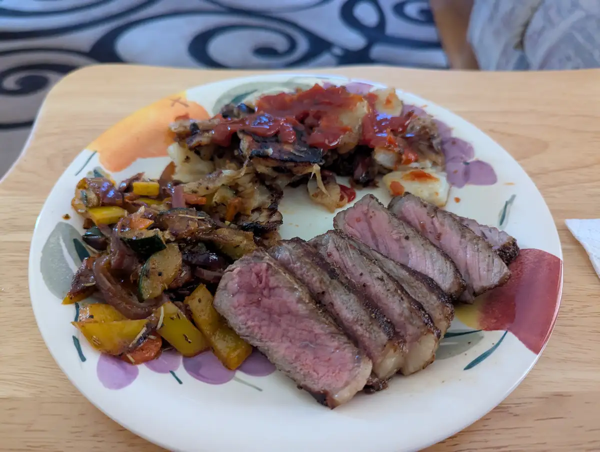 A plate of sliced medium-rare steak, roasted vegetables, and a side of what appears to be hash browns or potatoes topped…
