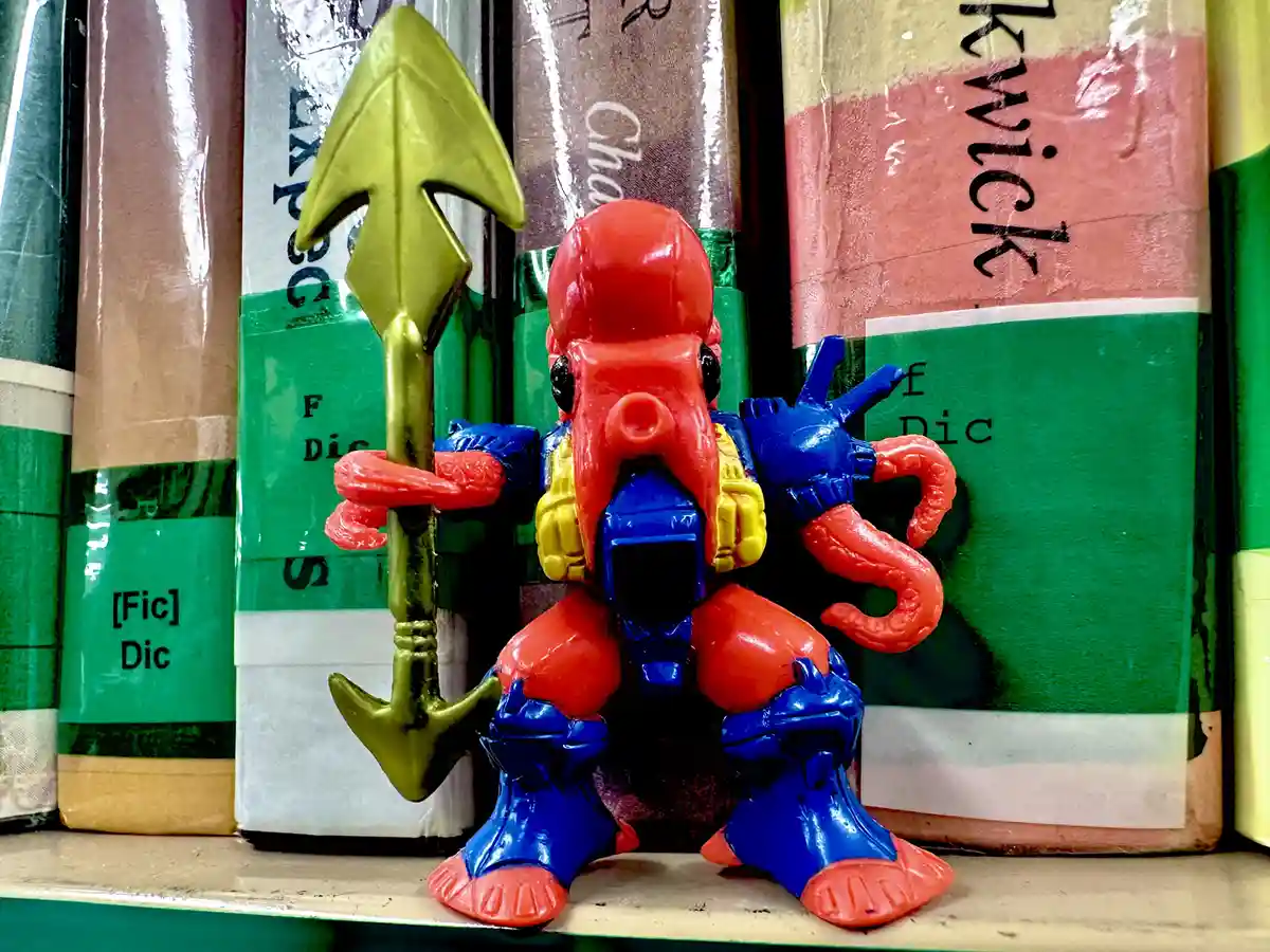 A red and blue plastic toy figure resembling an octopus warrior holding a gold spear stands on a shelf among wrapped books…