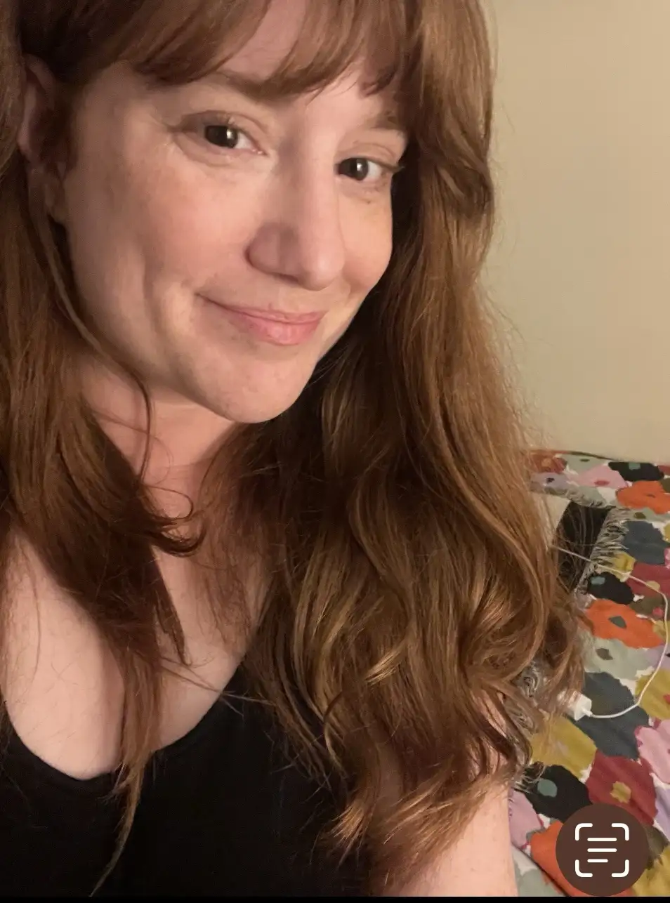 A close-up selfie of a woman with long, wavy brown hair and a subtle, knowing smile, wearing a black top. Behind her is a…