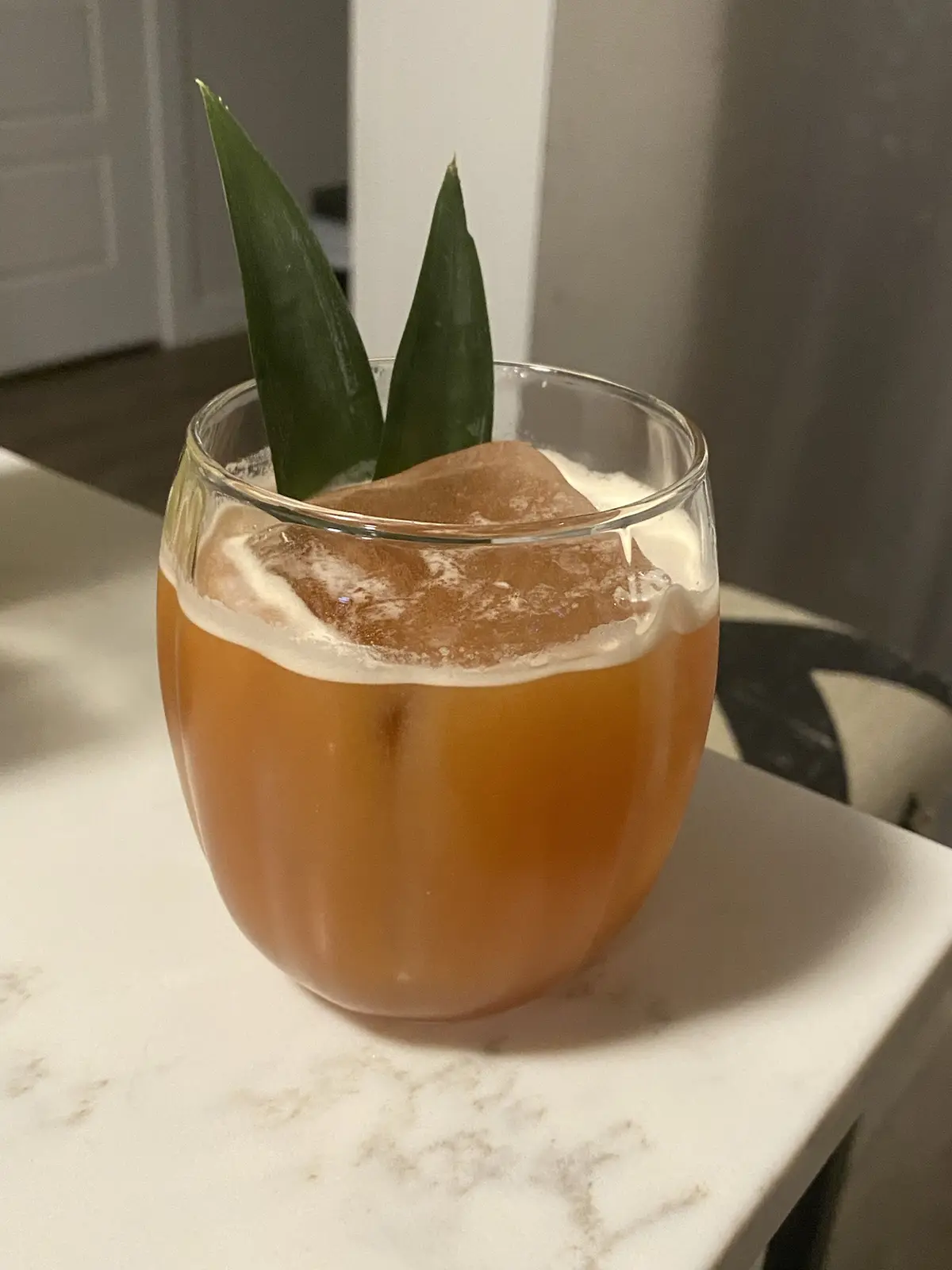 A close-up photo shows an amber-colored cocktail in a clear glass, topped with a large, rounded ice cube and two green…