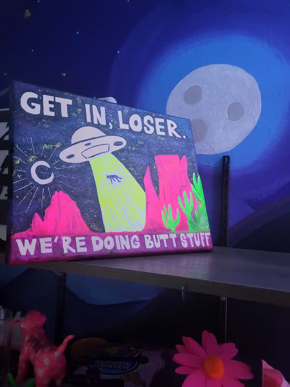A hand-painted canvas depicts a UFO abducting a person with a beam over pink desert rock formations and cacti, under a…
