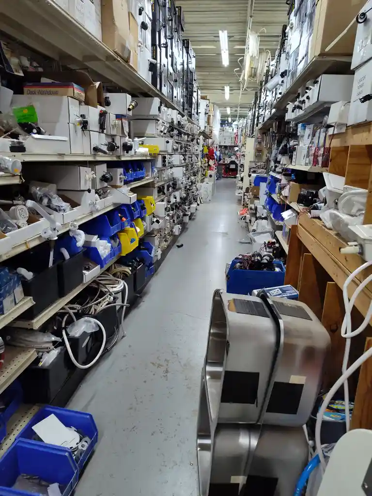 A long, narrow aisle in a hardware store is packed with shelves on both sides overflowing with boxes, bins, and plumbing…
