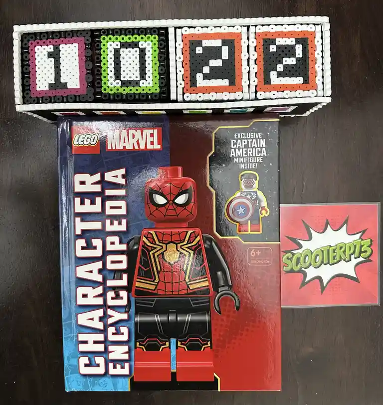 A LEGO Marvel Character Encyclopedia box featuring a Spider-Man minifigure is displayed on a dark wood surface. Above it, a…