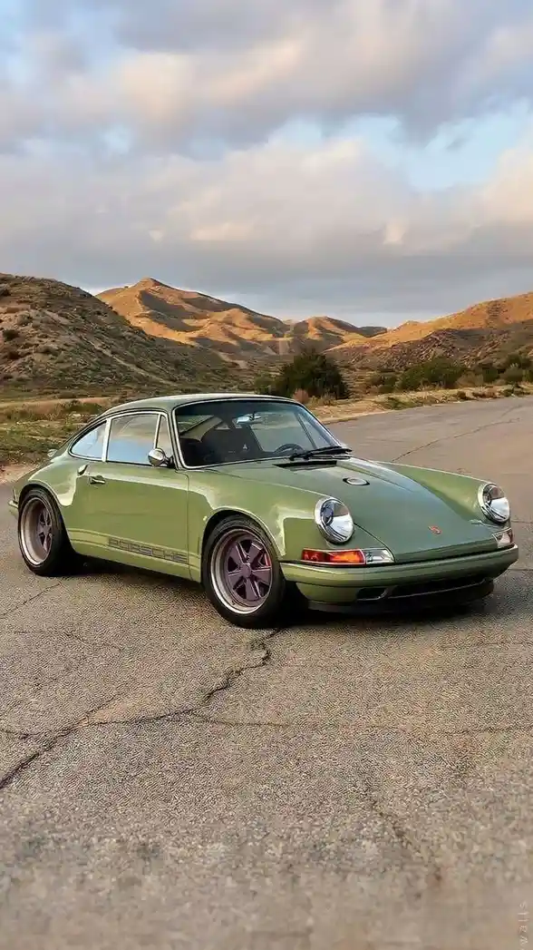 a light green classic porsche 911 is parked on a cracked asphalt road with rolling hills and a cloudy sky in the background…