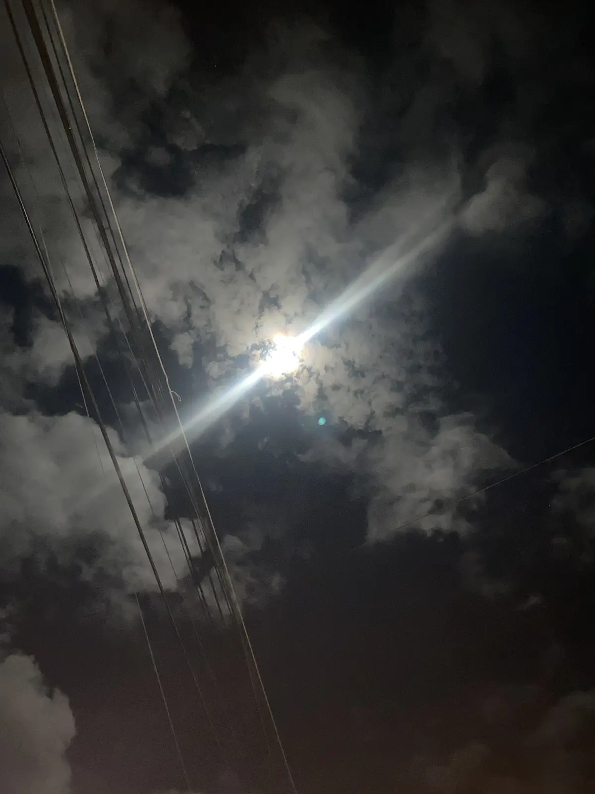 A night sky photo showing a bright moon partially obscured by clouds, with power lines running diagonally across the frame…