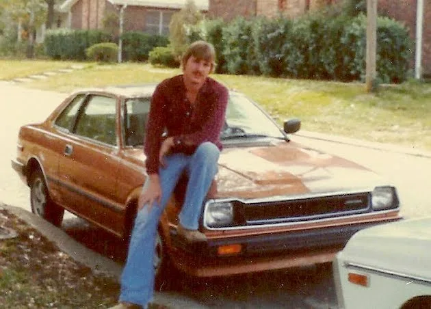 A man with a mustache and long hair sits casually on the hood of a rusty brown 1980s-style coupe in a suburban driveway. The…