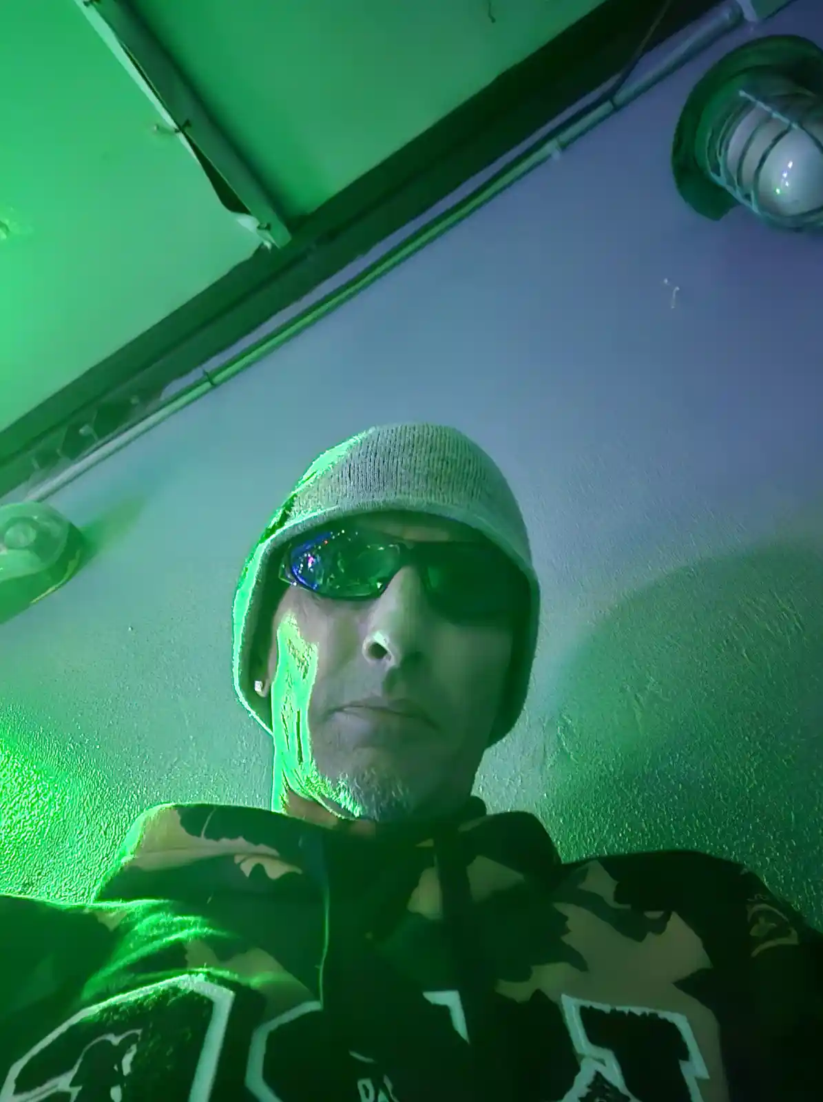 A man wearing a beanie and sunglasses looks directly at the camera under intense green lighting, with the text 'i'm not a…